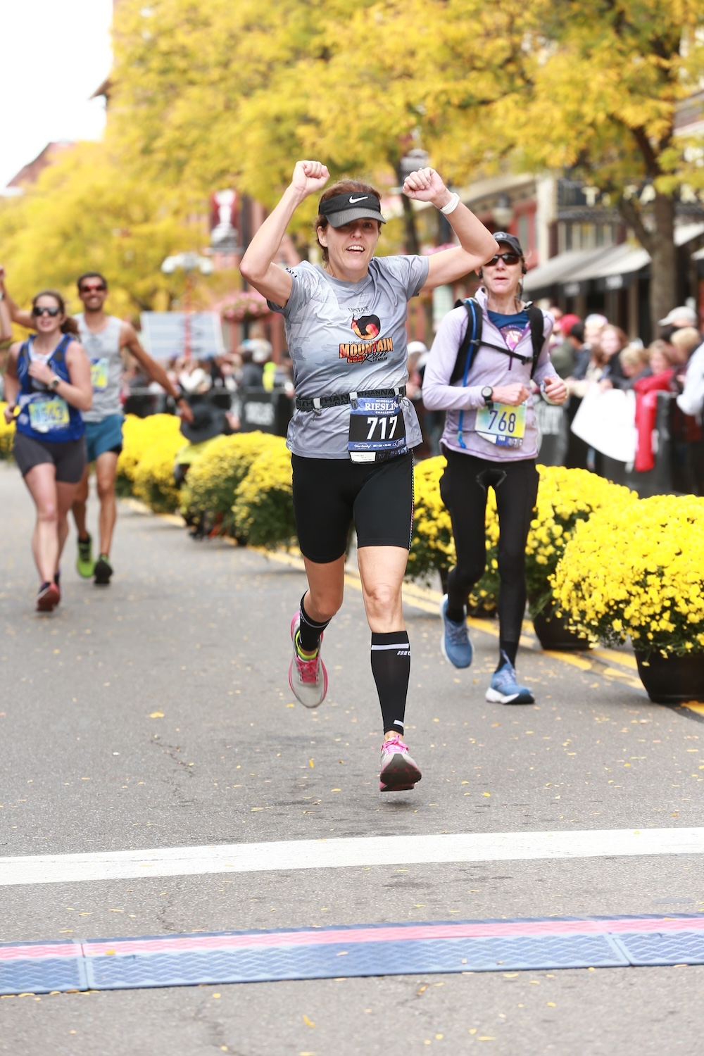 Runner with bib number 717 crosses a finish line with arms raised, lined by yellow flowers and other racers.