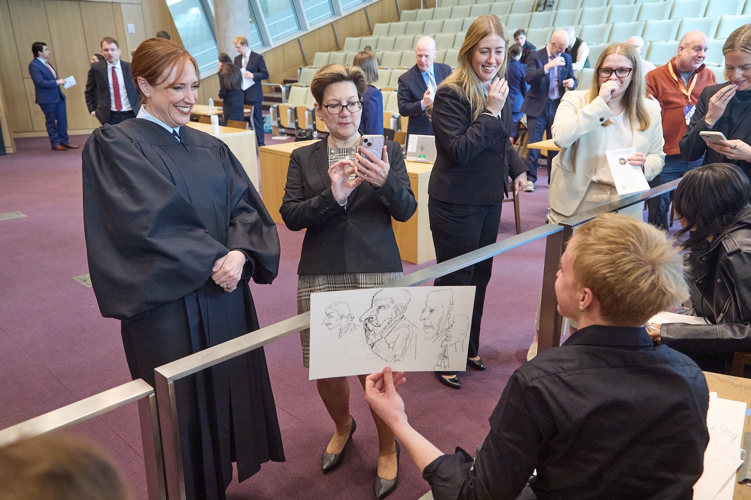 Person in a robe and others standing at a courtroom railing, looking at a hand-drawn courtroom sketch held up by an artist