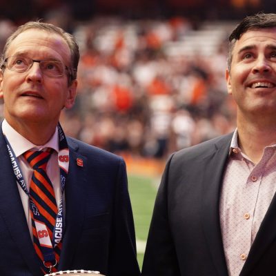 Two people in suits standing on a football field, one holding a Syracuse “S” football, with a stadium crowd behind them and a “Beat Navy” pin visible on a lapel.