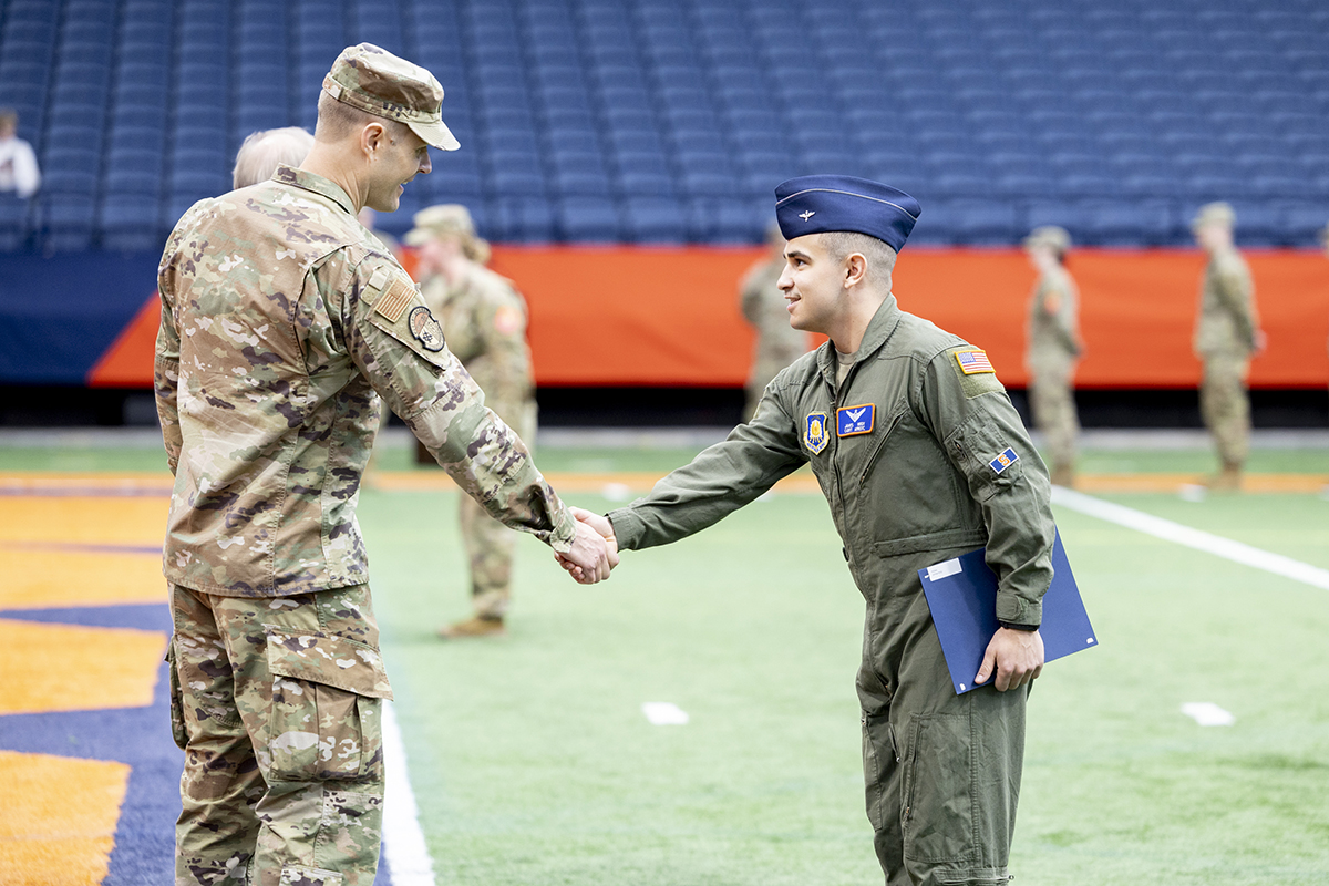 ROTC cadet shakes hands with a military officer during a formal recognition ceremony on an indoor field.