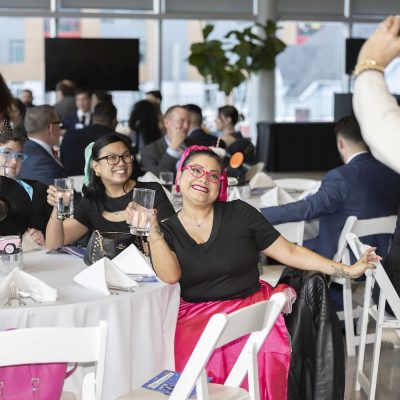 Guests seated at a banquet table raise glasses for a toast while another attendee takes a photo in a bright event space.