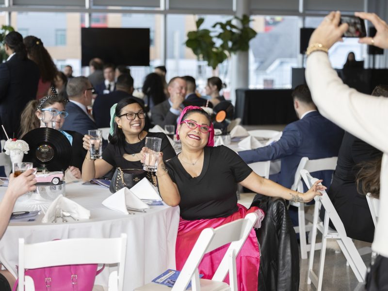 Guests seated at a banquet table raise glasses for a toast while another attendee takes a photo in a bright event space.