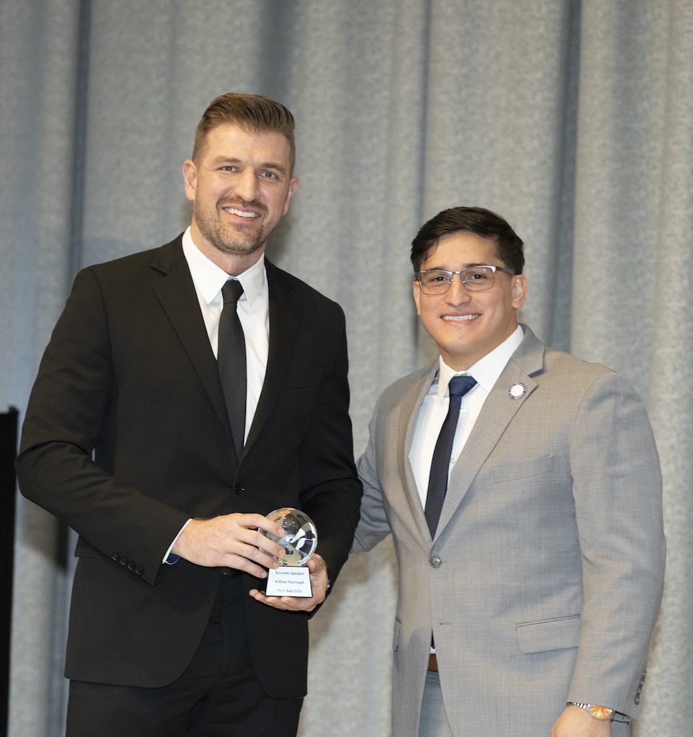Two people in suits stand together holding a glass award in front of a curtain backdrop.