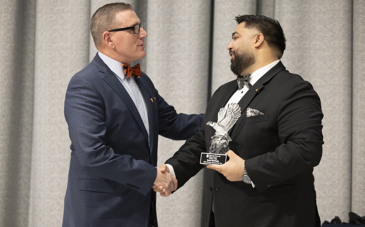 Two people in formal suits shake hands while one holds a glass award in front of a curtain backdrop.