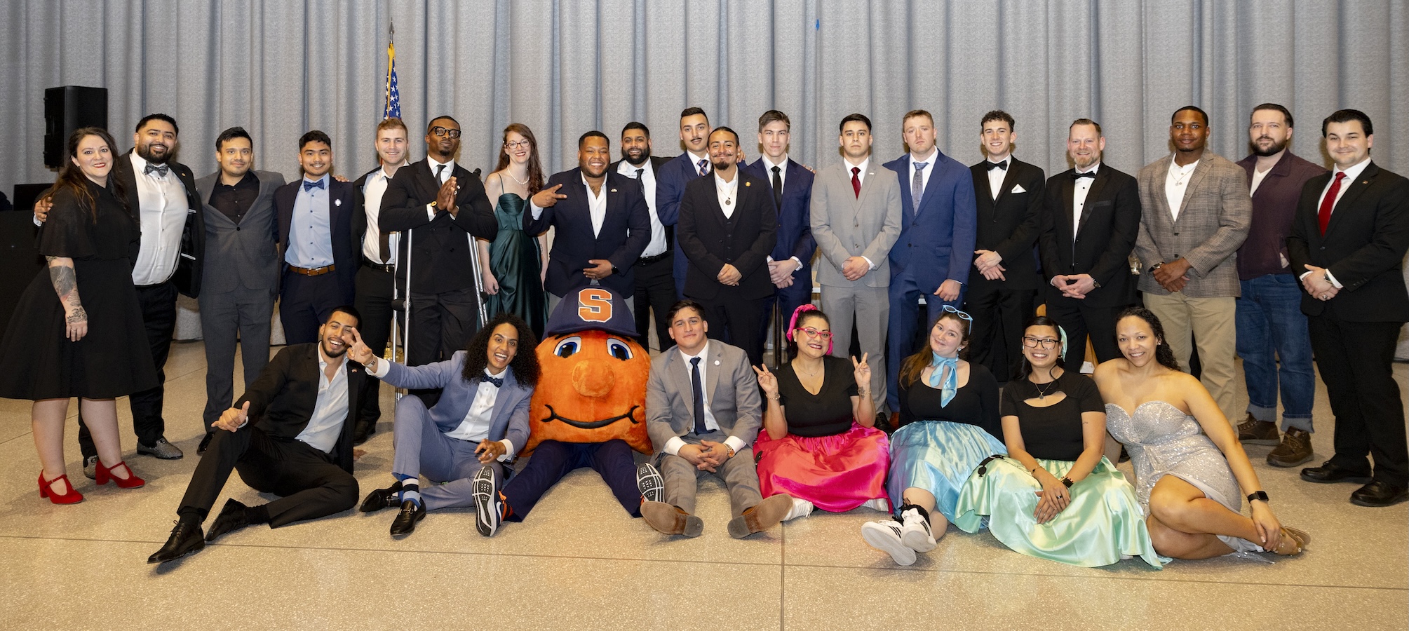 A large group in formal attire poses for a group photo on a stage, with an orange mascot seated front and center.