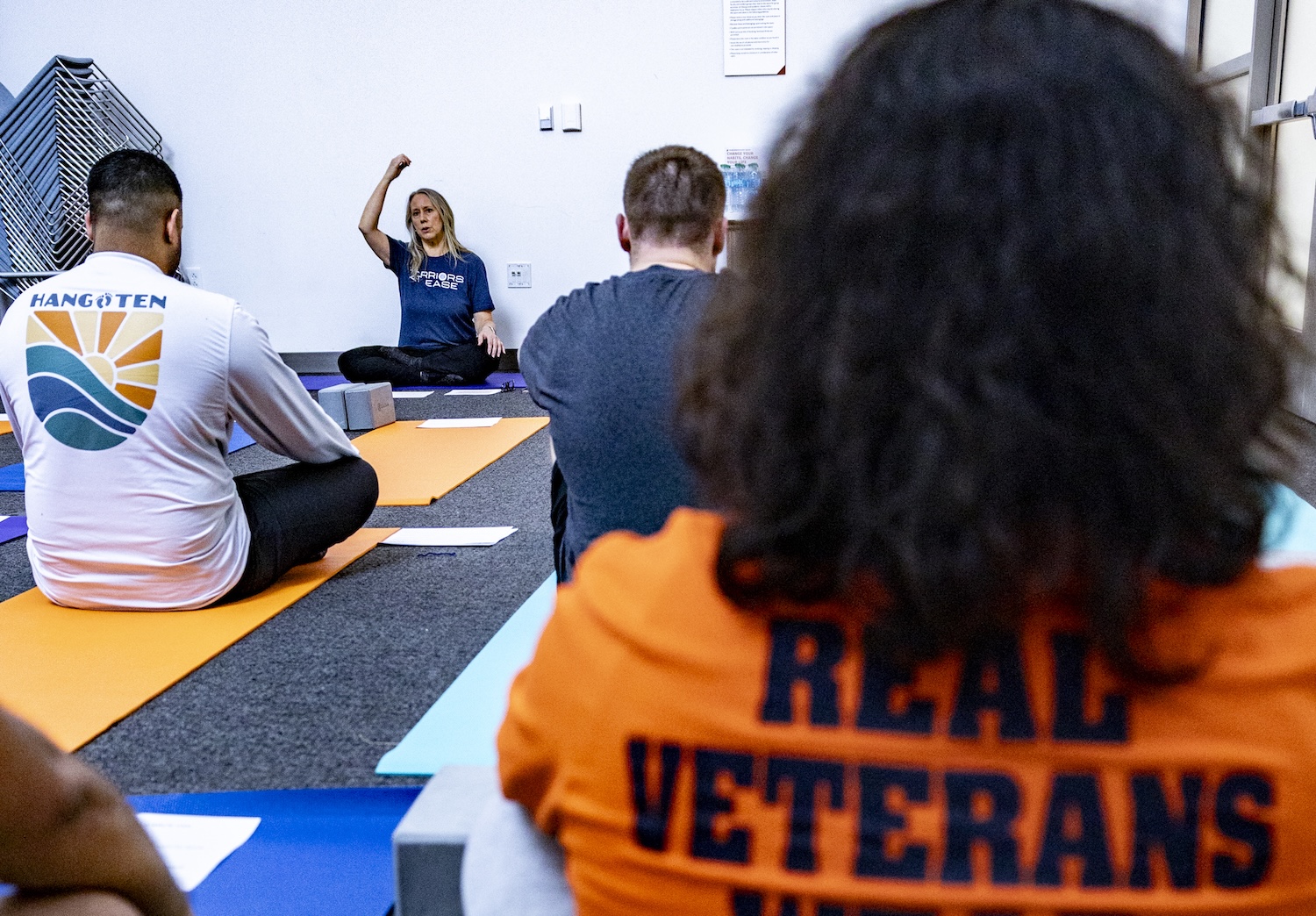 Instructor leading a group yoga class, with participants seated on mats in a studio, some wearing “REAL VETERANS” and “HangTen” shirts.