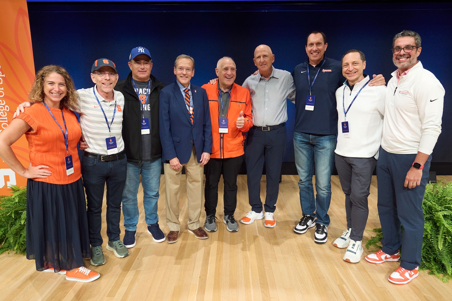 A group of people wearing name badges stands shoulder to shoulder on a stage, smiling for a group photo against a dark blue backdrop with orange accents.