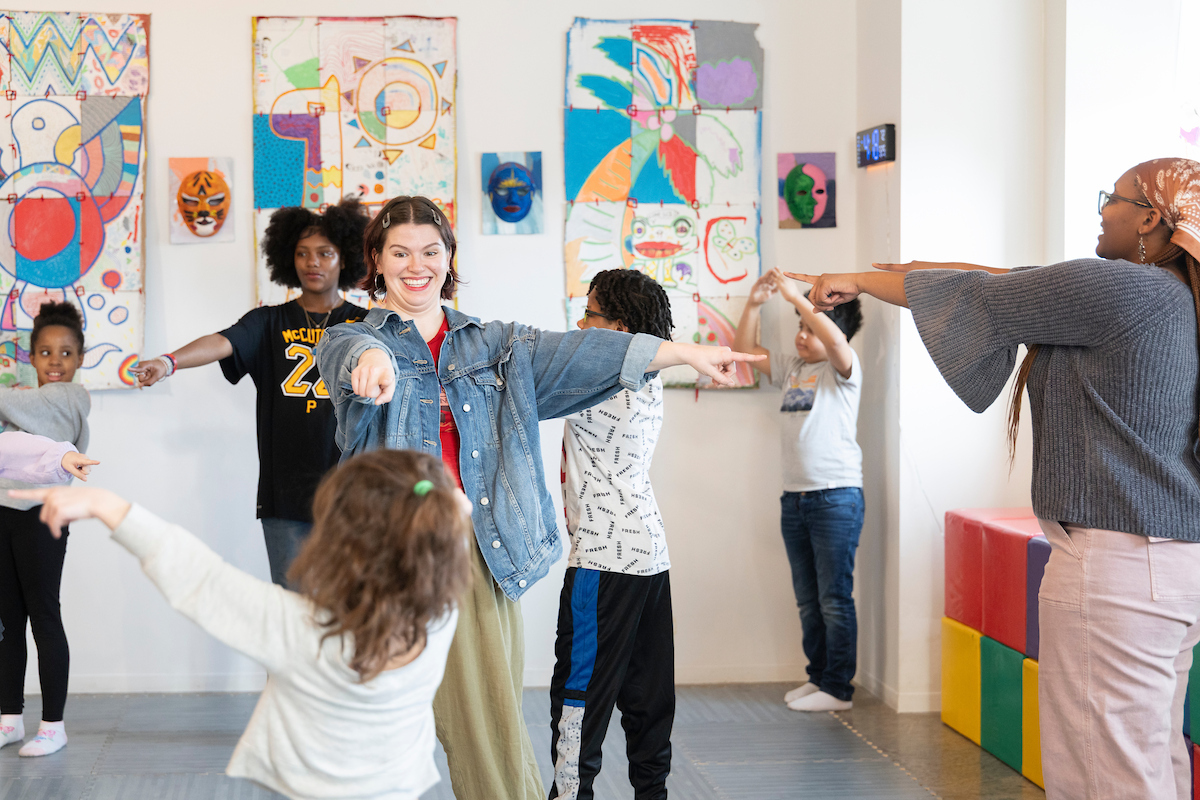 Group of children and young adults stretching and pointing together in a colorful classroom.
