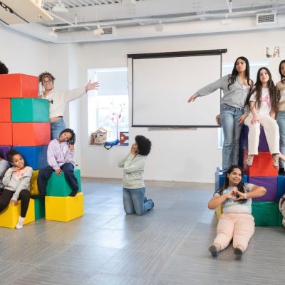 A large group of children and teens pose playfully in the La Casita Cultural Center, climbing on and arranging themselves around two towers of colorful foam blocks. Artwork lines the walls and a projection screen is visible in the background.