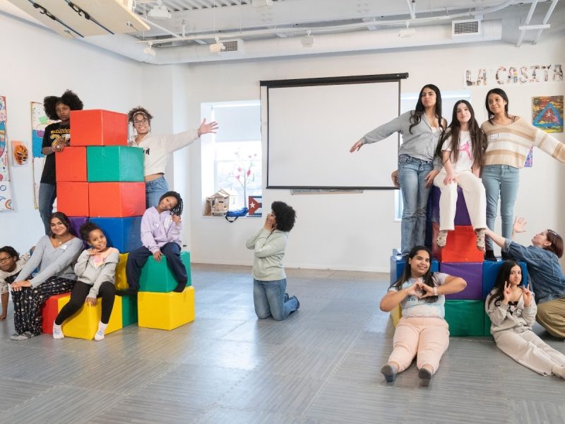 A large group of children and teens pose playfully in the La Casita Cultural Center, climbing on and arranging themselves around two towers of colorful foam blocks. Artwork lines the walls and a projection screen is visible in the background.