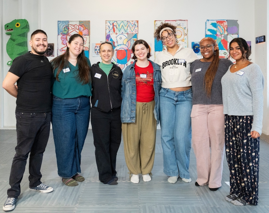Seven people smile for a group photo in an art-filled gallery space, with colorful student artwork and a green dinosaur sculpture displayed on the wall behind them. Several members of the group wear name tags.