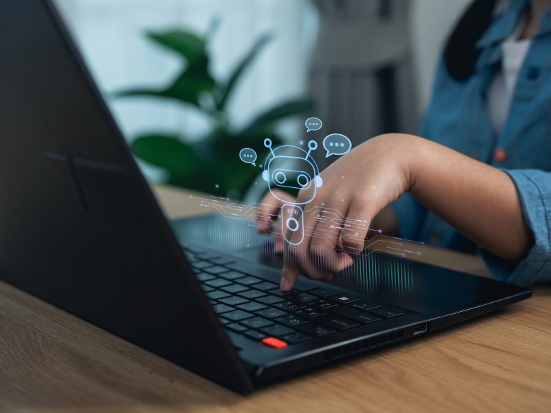 Hands typing on a laptop with a floating AI chatbot icon overlay above the keyboard.