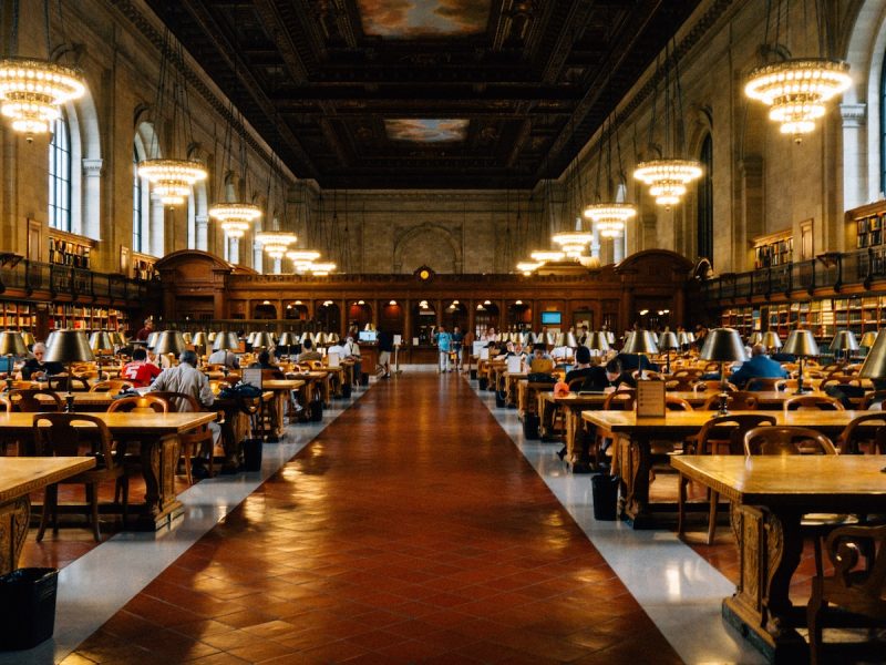 Grand library reading room with long wooden tables, green desk lamps, chandeliers, and readers seated beneath a high, ornate ceiling.