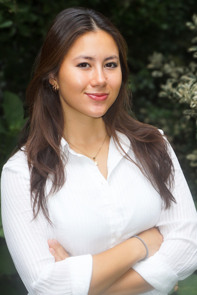 Portrait of a person standing outdoors with arms crossed, wearing a white blouse, with greenery in the background.