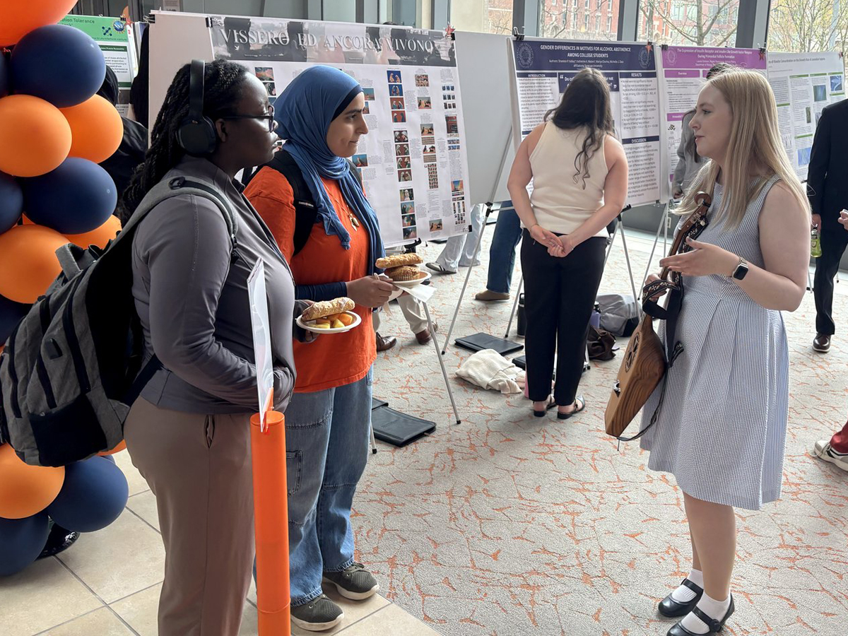 A student holding a panduri speaks with two attendees at an academic research poster session with orange and blue balloons.