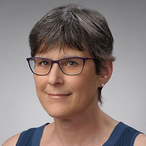 Professional headshot of a woman with short gray-brown hair wearing purple-framed glasses and a navy blue sleeveless top, photographed against a neutral gray background.
