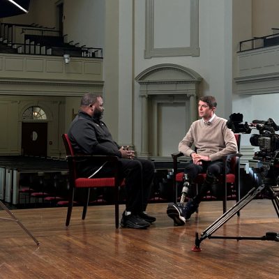 Two men sit facing each other in chairs inside a large chapel, engaged in conversation during a filmed interview. A camera on a tripod and studio lighting equipment are visible in the foreground, with rows of empty pews and ornate architectural details in the background.