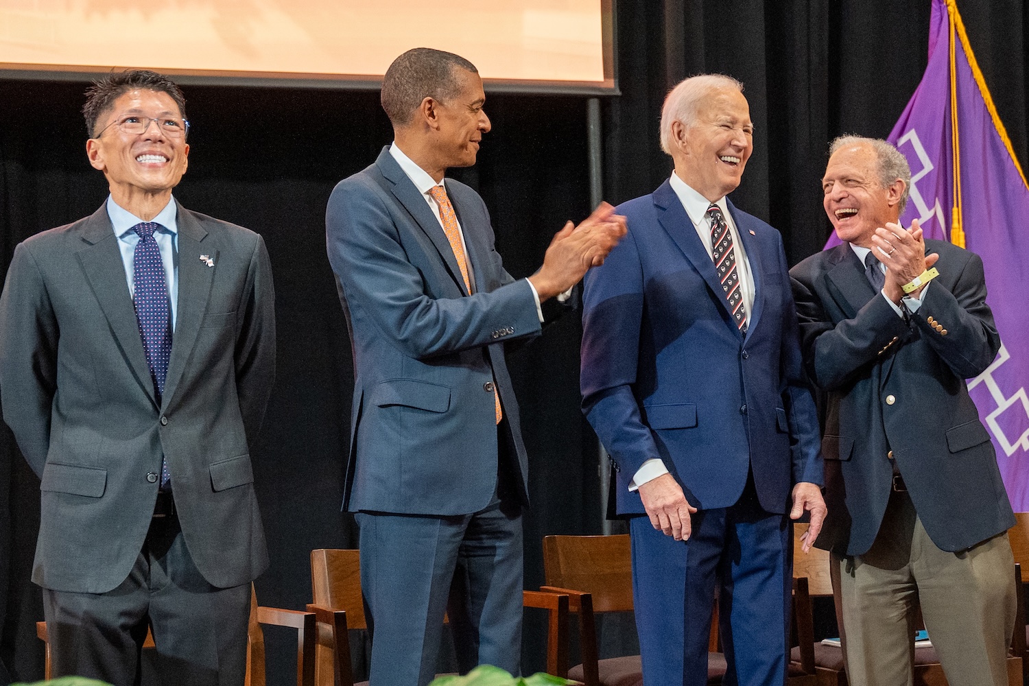 Four people in suits stand on a stage applauding, with chairs behind them.