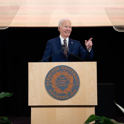 Speaker stands at a Syracuse University podium, gesturing during a speech, with teleprompters on either side.