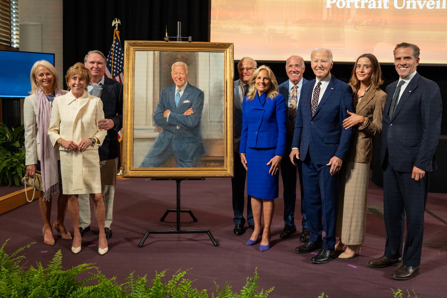 Group poses on a stage around a framed portrait on an easel, with an American flag and presentation screen in the background.