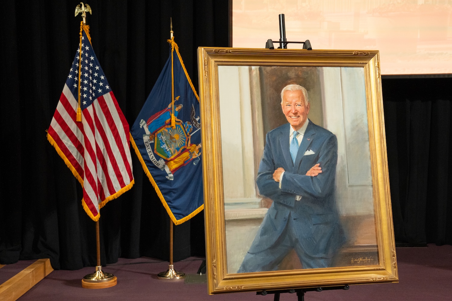 A painted portrait of former President Joe Biden in a gold frame, displayed on a stage beside U.S. and New York state flags.