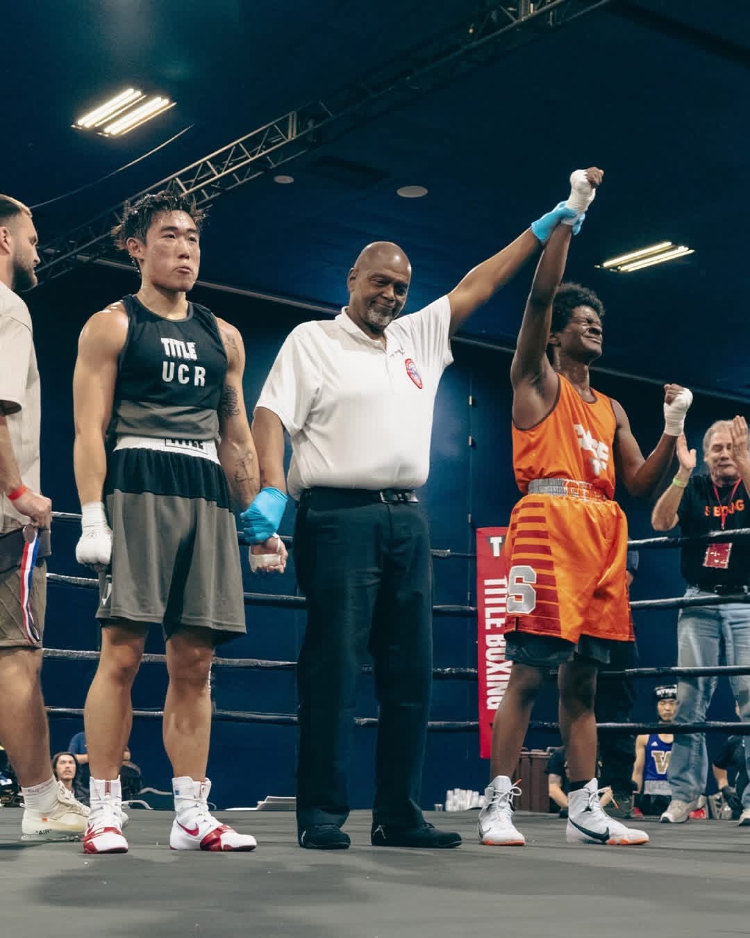 A referee raises the arm of a boxer in orange during a match in a boxing ring, with the opposing boxer standing nearby.