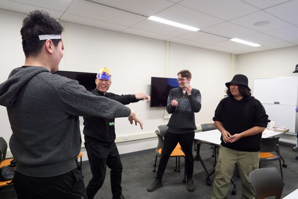 A group of people in a room having fun. They are engaged in an activity with two wearing playful paper crowns. The room has white walls, two flat screen TVs, and a whiteboard. The atmosphere is casual and lively.