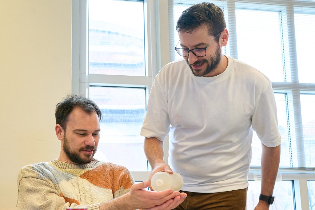 Two people examine a small white round object together in a bright, windowed room. One person is seated and wearing a patterned sweater; the other is standing and wearing a white T-shirt, holding the object.
