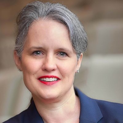 Head-and-shoulders portrait of a woman with short gray hair, red lipstick and a navy blazer, smiling against a blurred auditorium background.