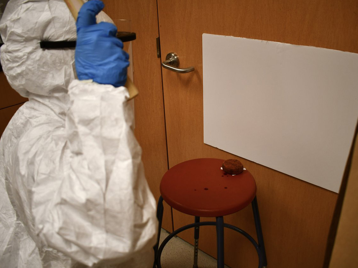 A student in a white protective suit and blue gloves collects evidence from a door handle at a simulated crime scene.