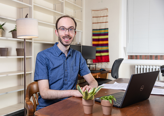 A person sits at a wooden desk with a laptop and papers, facing the camera in a home office with shelves, plants, and a colorful wall hanging in the background.