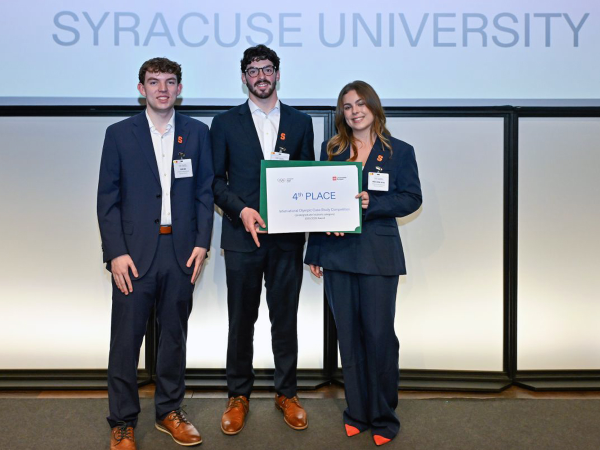 Three students stand on a stage at the Olympic Museum with the words Syracuse University on a screen behind them.