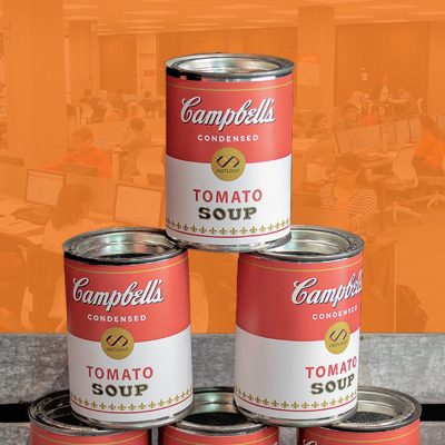 Stack of Campbell’s condensed tomato soup cans arranged in a pyramid against an orange background.
