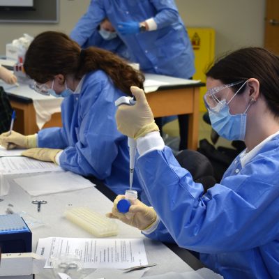 Students in blue gowns, masks and gloves conduct lab work, one using a pipette to transfer liquid into a vial.
