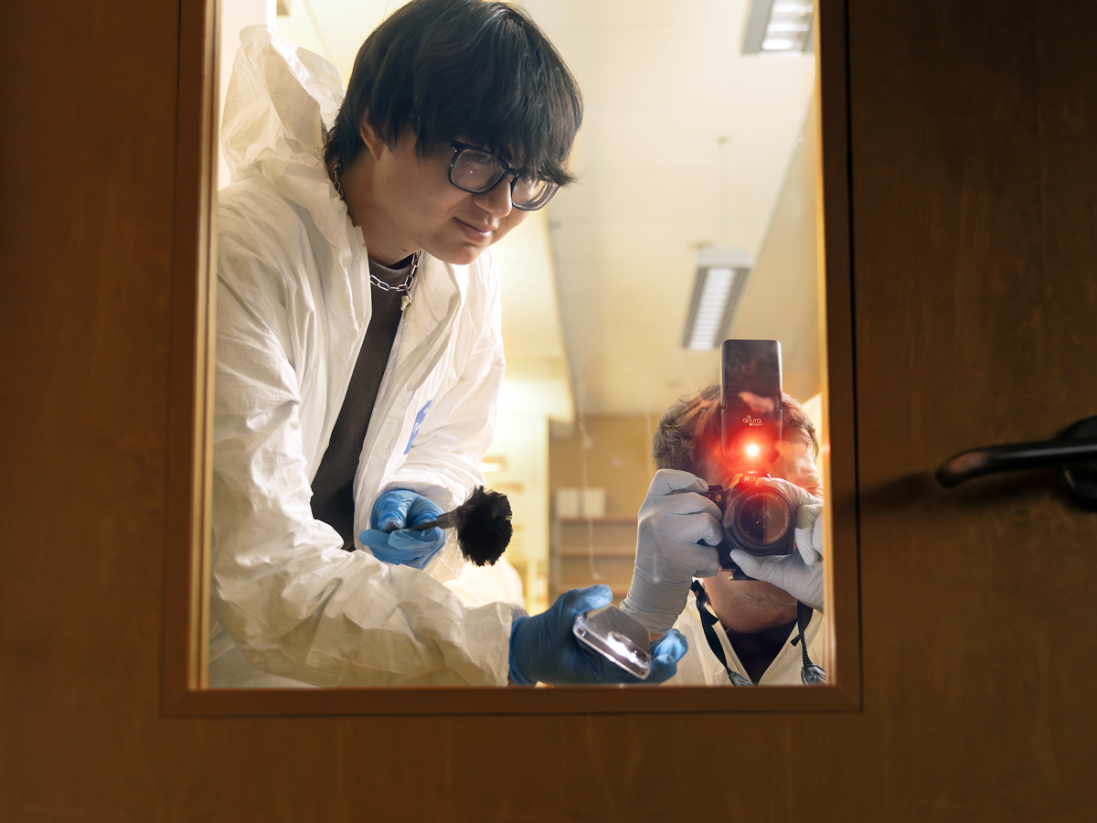 A student in a white protective suit dusts for fingerprints on a door while a photographer captures the scene.