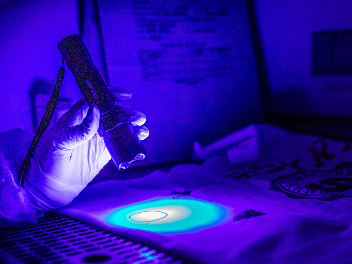 A gloved hand holds a UV flashlight illuminating a circular glowing mark on fabric in a darkened room.