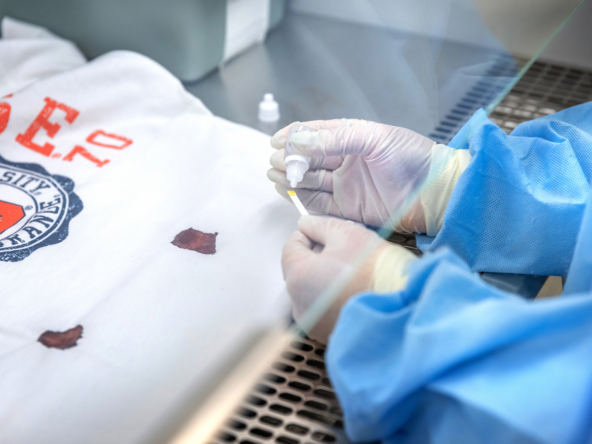A gloved researcher in a blue gown applies solution to a test strip near blood stains on a white shirt.