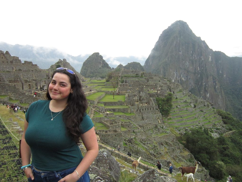 A Syracuse Abroad student poses at Machu Picchu in Peru, with the ancient Inca stone terraces, ruins and Huayna Picchu mountain visible behind her under an overcast sky.