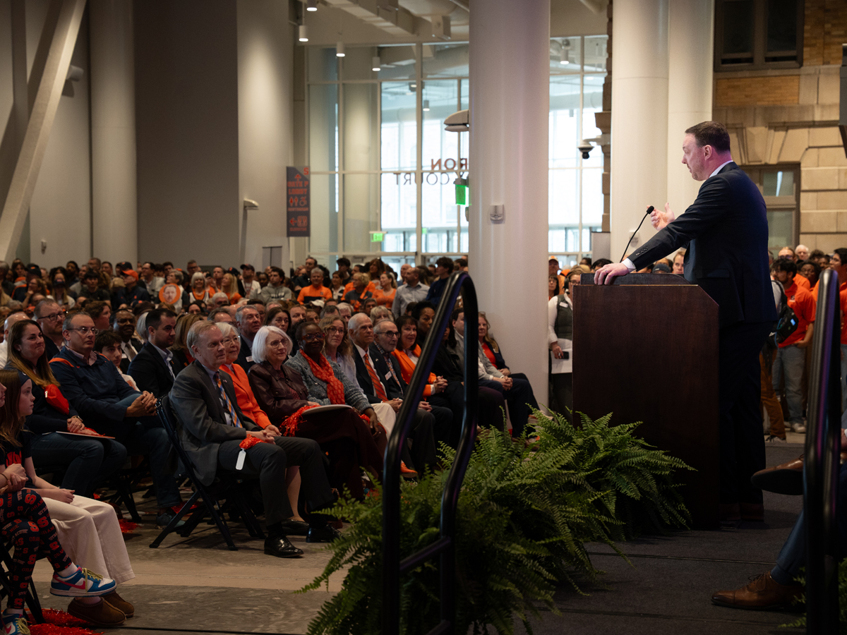 A man speaks at a podium before a large crowd of orange-clad attendees gathered in Miron Victory Court.
