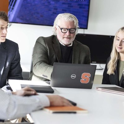 A small group sits around a table reviewing a laptop during a collaborative meeting in a modern office setting.