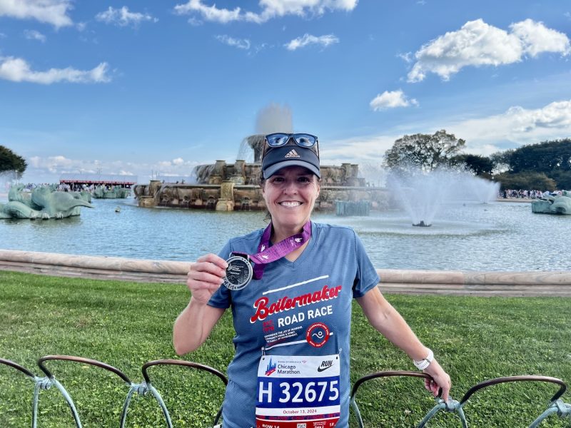Runner holding a finisher medal in front of Buckingham Fountain, wearing a “Buttermaker Road Race” shirt and Chicago Marathon bib.