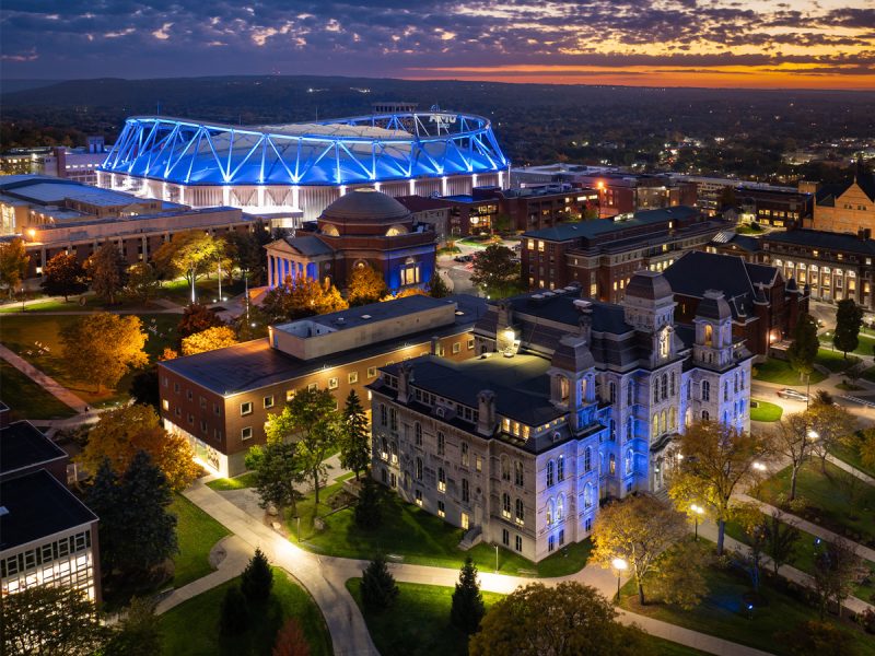 Aerial view of a university campus at dusk with historic academic buildings and a large stadium illuminated in blue light.