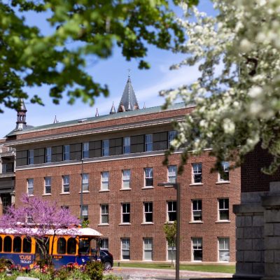 Brick campus building in spring with blooming trees and a colorful trolley bus passing in the foreground.