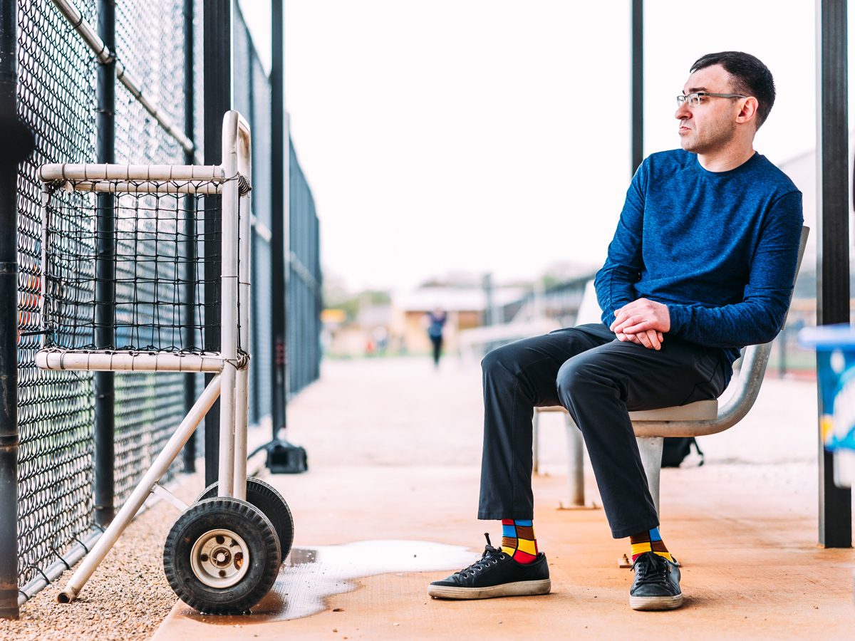A broadcaster sits on a bench in a baseball park wearing colorful patterned socks.
