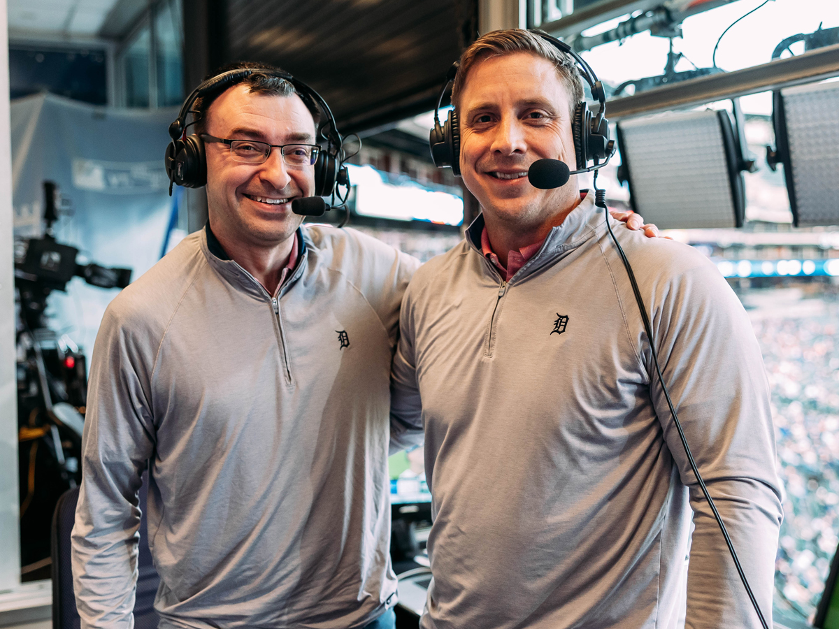 Two sportscasters smile while wearing Detroit Tigers gear inside the broadcast booth.