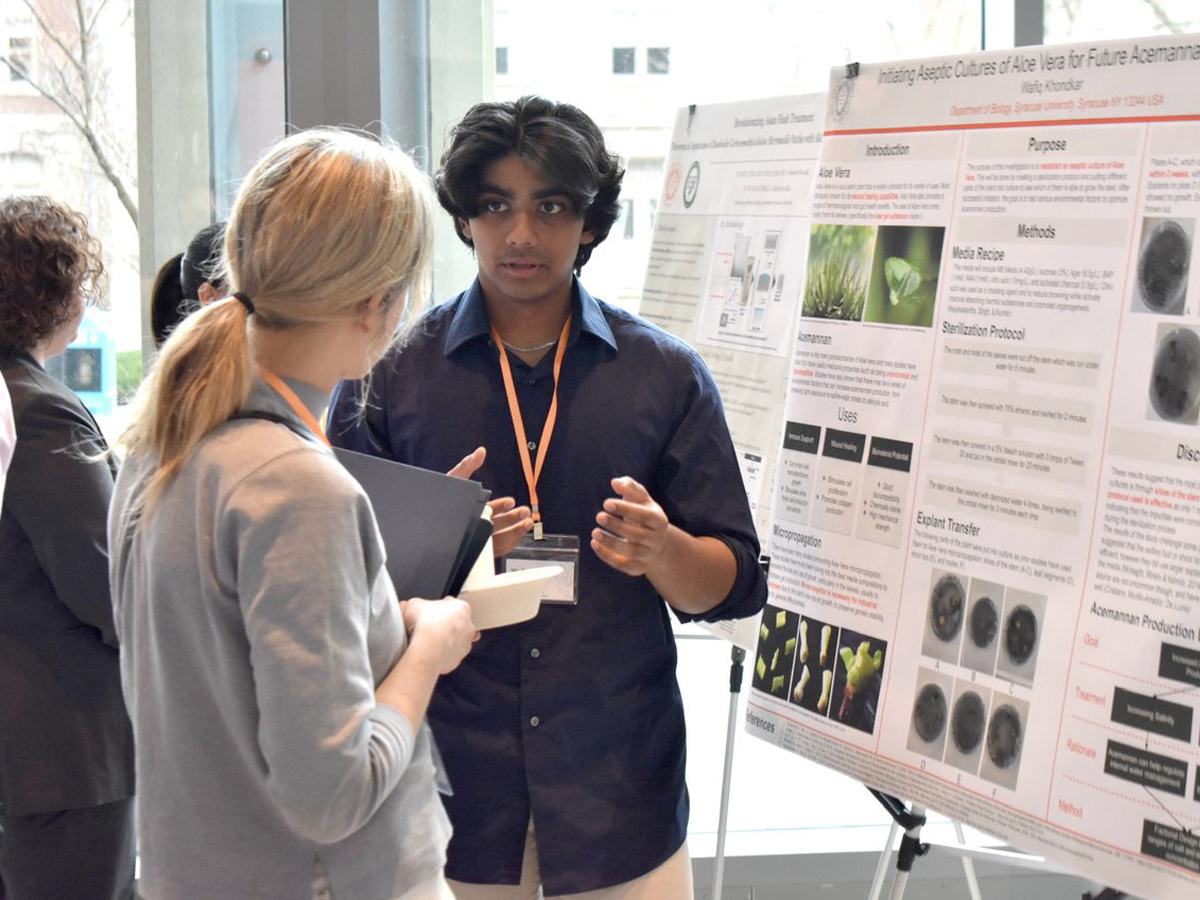 A student presents his research poster on aloe vera aseptic cultures to an attendee at an academic conference.