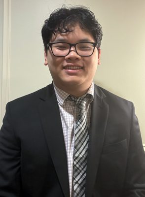 Smiling person wearing glasses, a suit jacket, and patterned tie against a neutral background