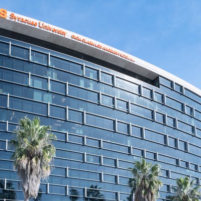 A modern glass office building with Syracuse University's Dick Clark Los Angeles Program signage in orange, framed by palm trees and a blue sky.