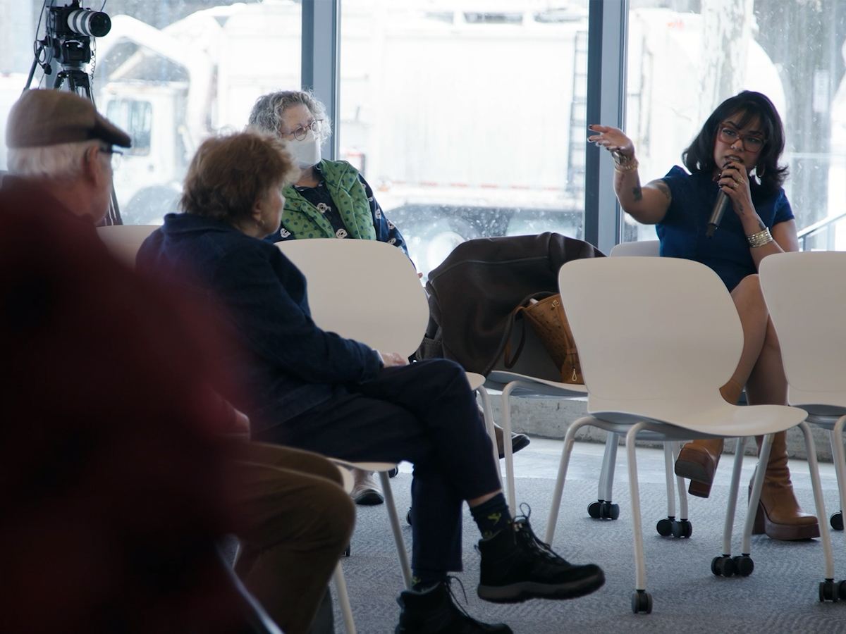 A woman speaks to a small seated audience during an indoor discussion event near large windows.