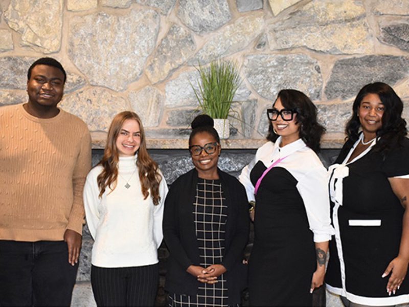 Five people smile together in front of a stone wall at an indoor event.